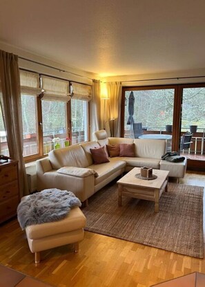 Ein gemütliches Wohnzimmer mit einer Couch, einem Hocker und einem Couchtisch, großen Fenstern mit Vorhängen und Blick ins Freie.