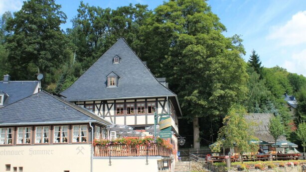 Un bâtiment traditionnel à colombages, doté d’un balcon, entouré d’une végétation luxuriante et d’arbres.