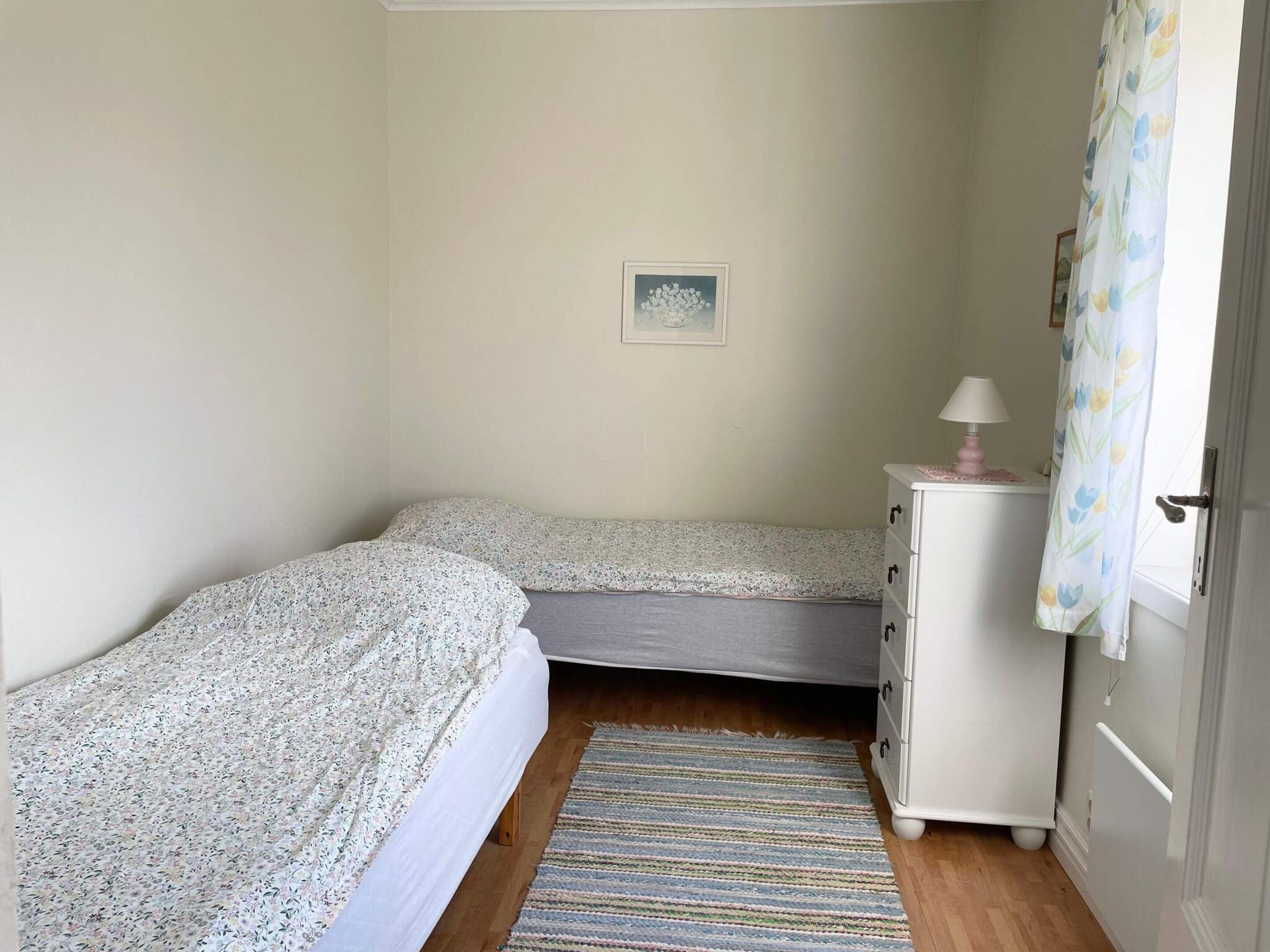 Quarto com duas camas de solteiro, mesa de cabeceira com abajur, cômoda pequena, tapete e um quadro na parede.
