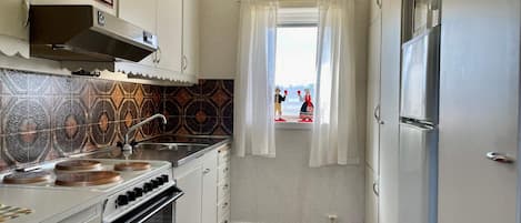 Cocina compacta con armarios blancos, estufa, horno y una ventana con cortinas.
