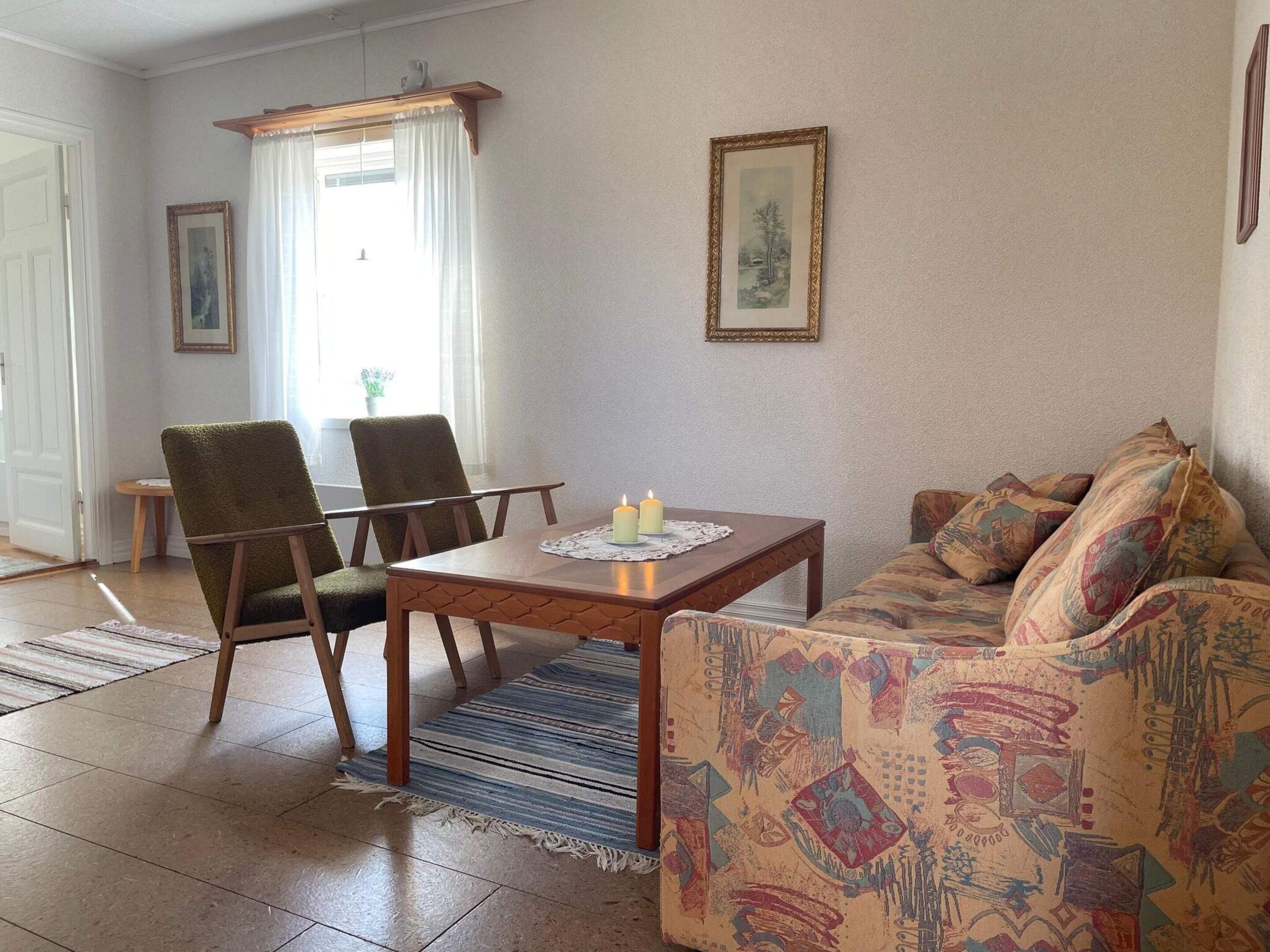 Una sala de estar con un sofá, dos sillones, una mesa con una vela y una ventana con cortinas.