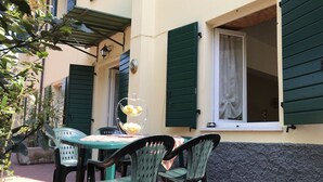 Une terrasse extérieure avec une table et des chaises, un panier de fruits en forme de cœur et une fenêtre avec des volets verts.
