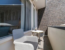 Een balkon met een rieten stoel, een klein rond tafeltje en uitzicht op een bakstenen muur.