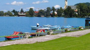 Vista de un lago con barcos amarrados a un camino de piedra, una iglesia con una alta aguja y edificios residenciales al fondo.