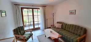 A room with a green patterned sofa, a wooden chair, a table with a white cloth, a radiator, and a window with curtains. - Ferienwohnung im Haus Schoenbrunn (Bayrischzell)