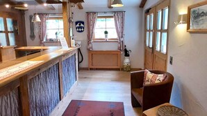 Un área de recepción con mostrador de madera, una mesa de recepción, una silla con cojín, una ventana con cortinas y una puerta.