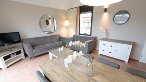 Een moderne woonkamer met een grijze bank, een houten eettafel met een vaas witte bloemen, een flatscreen televisie en een wit kastje.