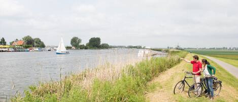 A riverside path with a sailboat in the distance, grassy banks, and a cyclist pointing towards the water.