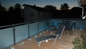 A wooden deck with a glass balustrade, outdoor seating, and a pool area at dusk. - Nice Apartment in Himmelkron (Himmelkron)
