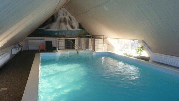 An indoor swimming pool with a waterfall feature, surrounded by a white ceiling and walls, and a small seating area with a ladder leading to the pool. - Nice Apartment in Himmelkron (Himmelkron)