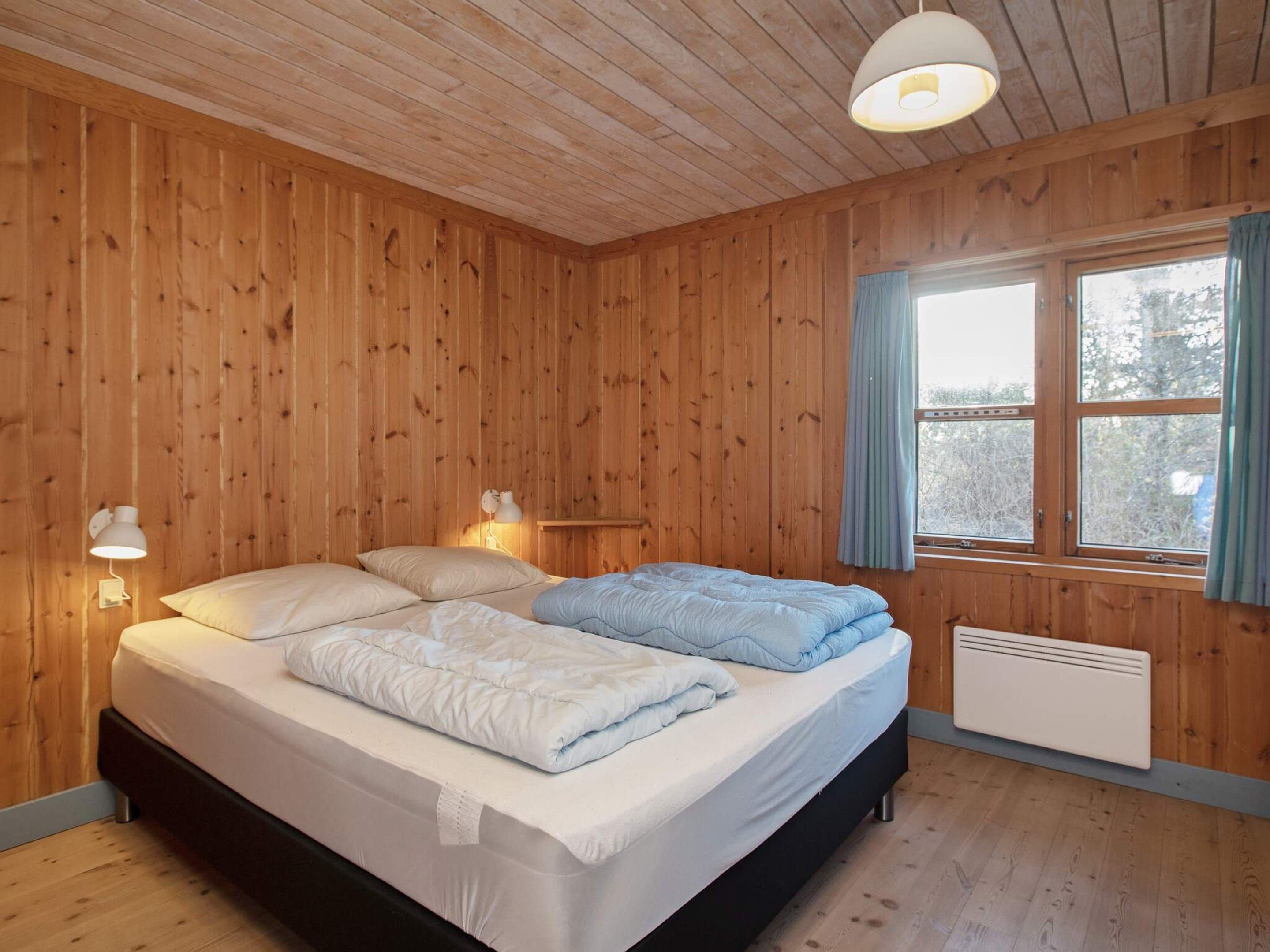 A wooden-paneled room with a bed, two bedside lamps, a window with curtains, and a radiator.
