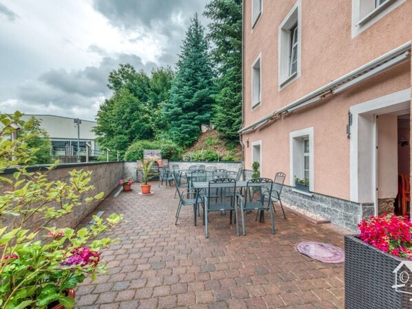 Un espace patio avec des sièges d’extérieur et un sol en briques, entouré de verdure et d’un bâtiment à la façade en pierre.