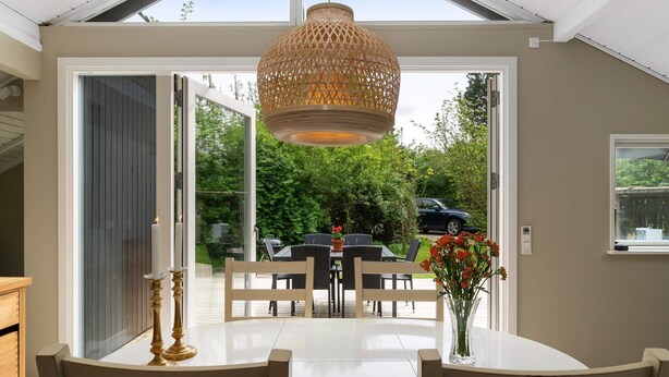 Un coin repas avec une table et des chaises blanches, un luminaire suspendu et une vue sur l’extérieur.