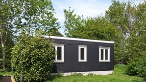 Un petit bâtiment moderne de couleur noire, doté de trois fenêtres, entouré d’une végétation luxuriante et agrémenté d’une terrasse en bois.