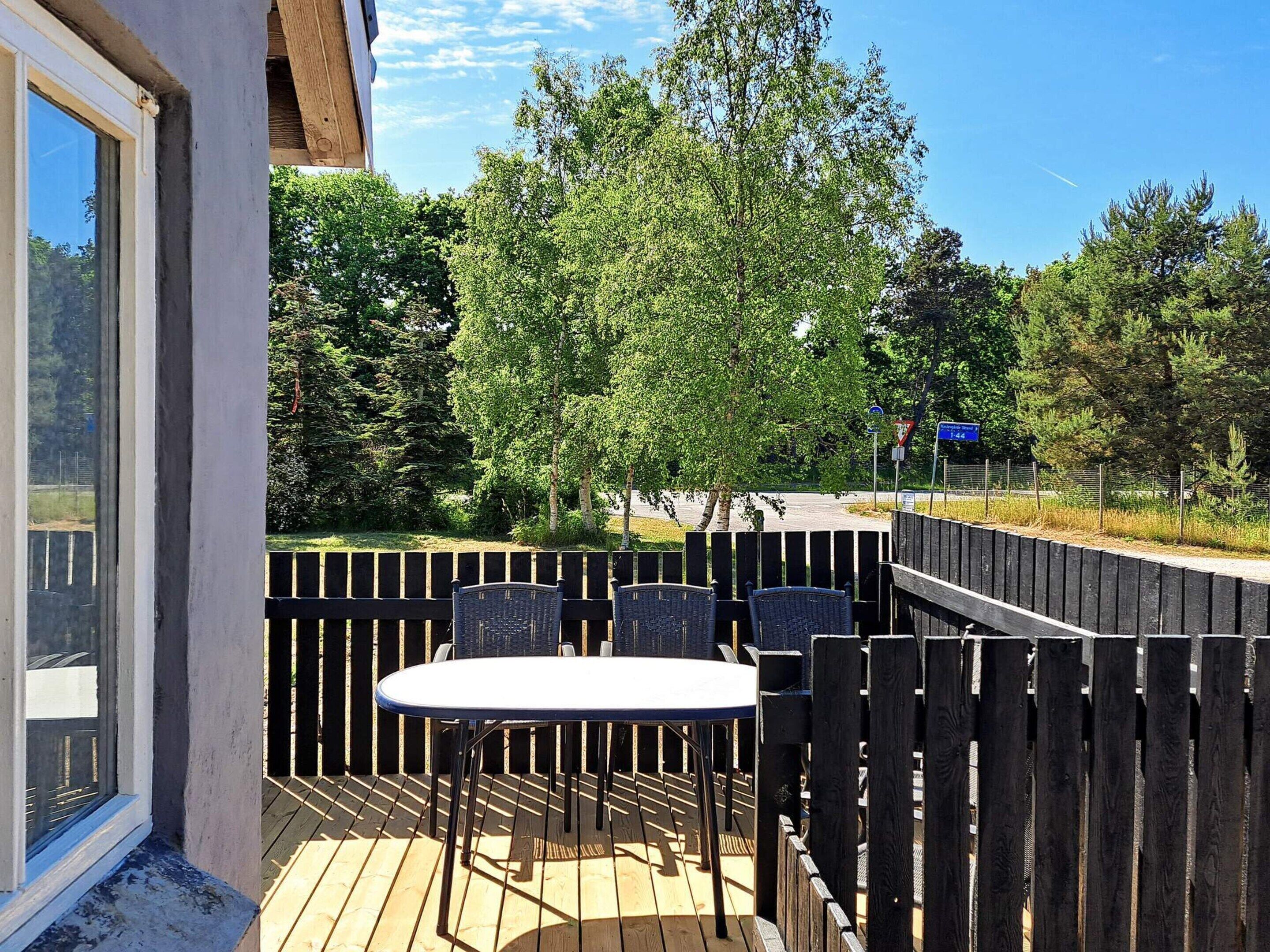 Una terraza exterior con una mesa y sillas, rodeada por una valla de madera, con árboles y una carretera al fondo.