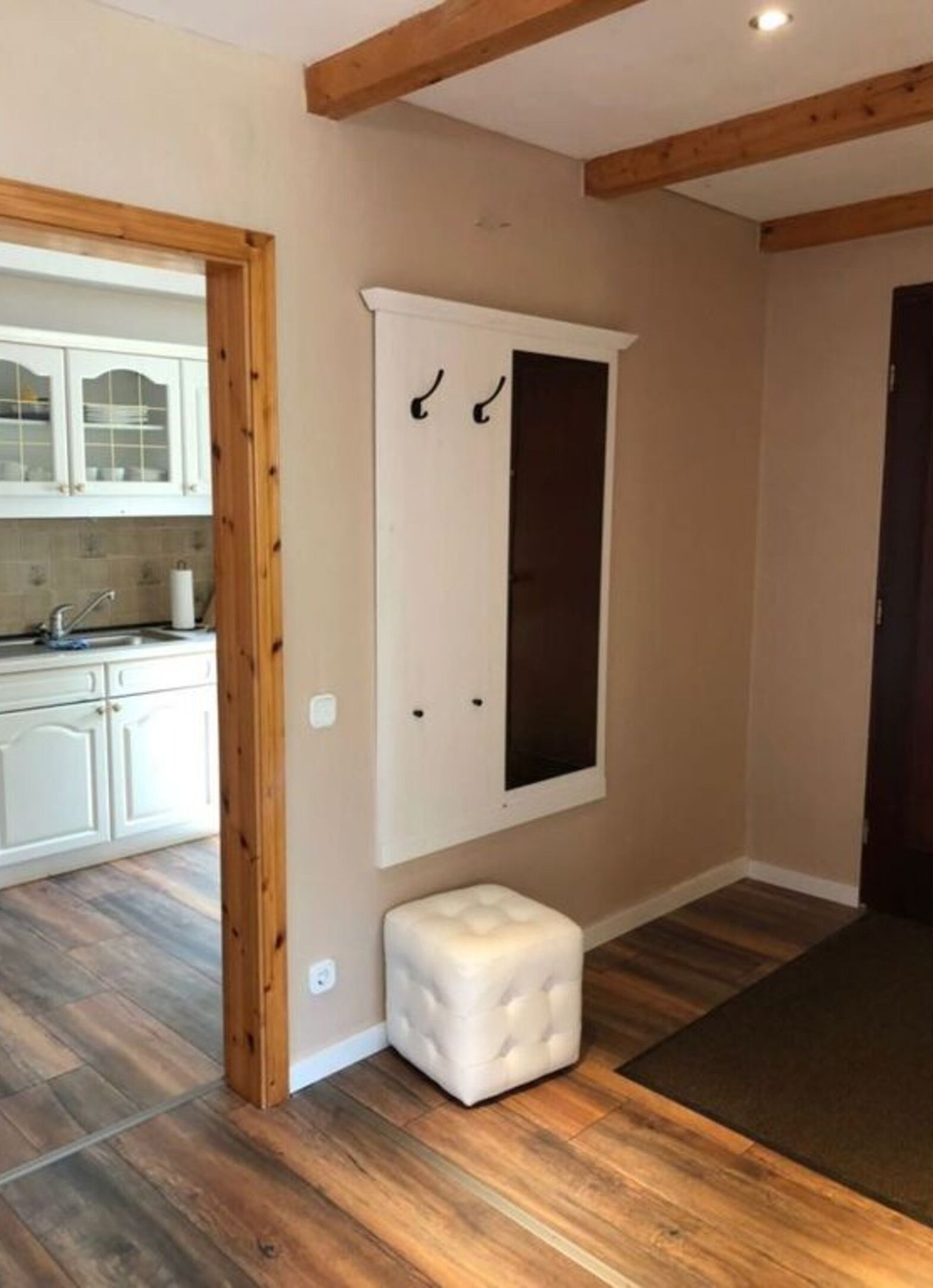 Una habitación con vigas de madera, un armario blanco, un pequeño otoman blanco y una cocina con armarios blancos y un lavamanos.