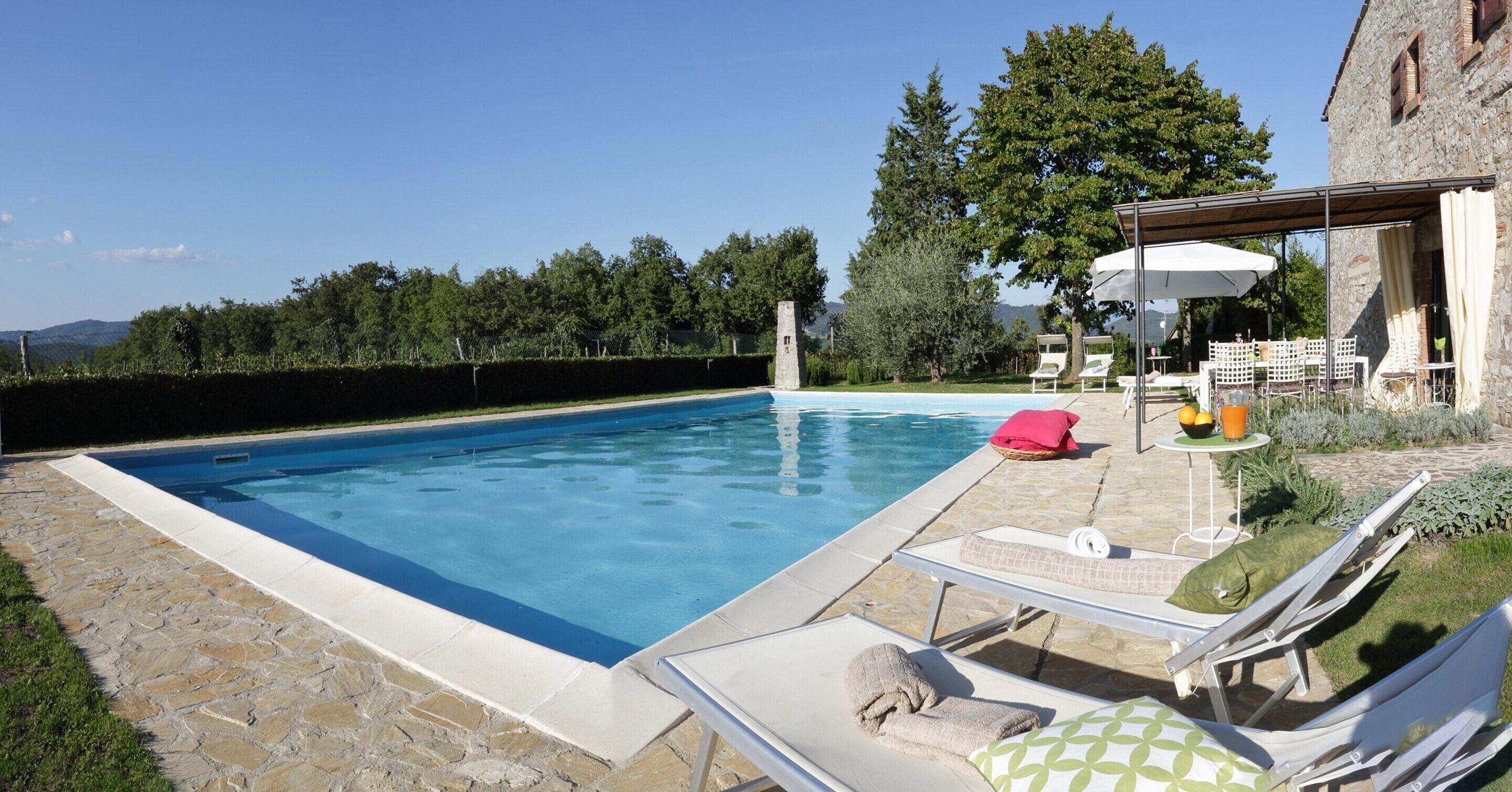 A swimming pool with lounge chairs and a table with umbrellas, surrounded by greenery and a stone building.
