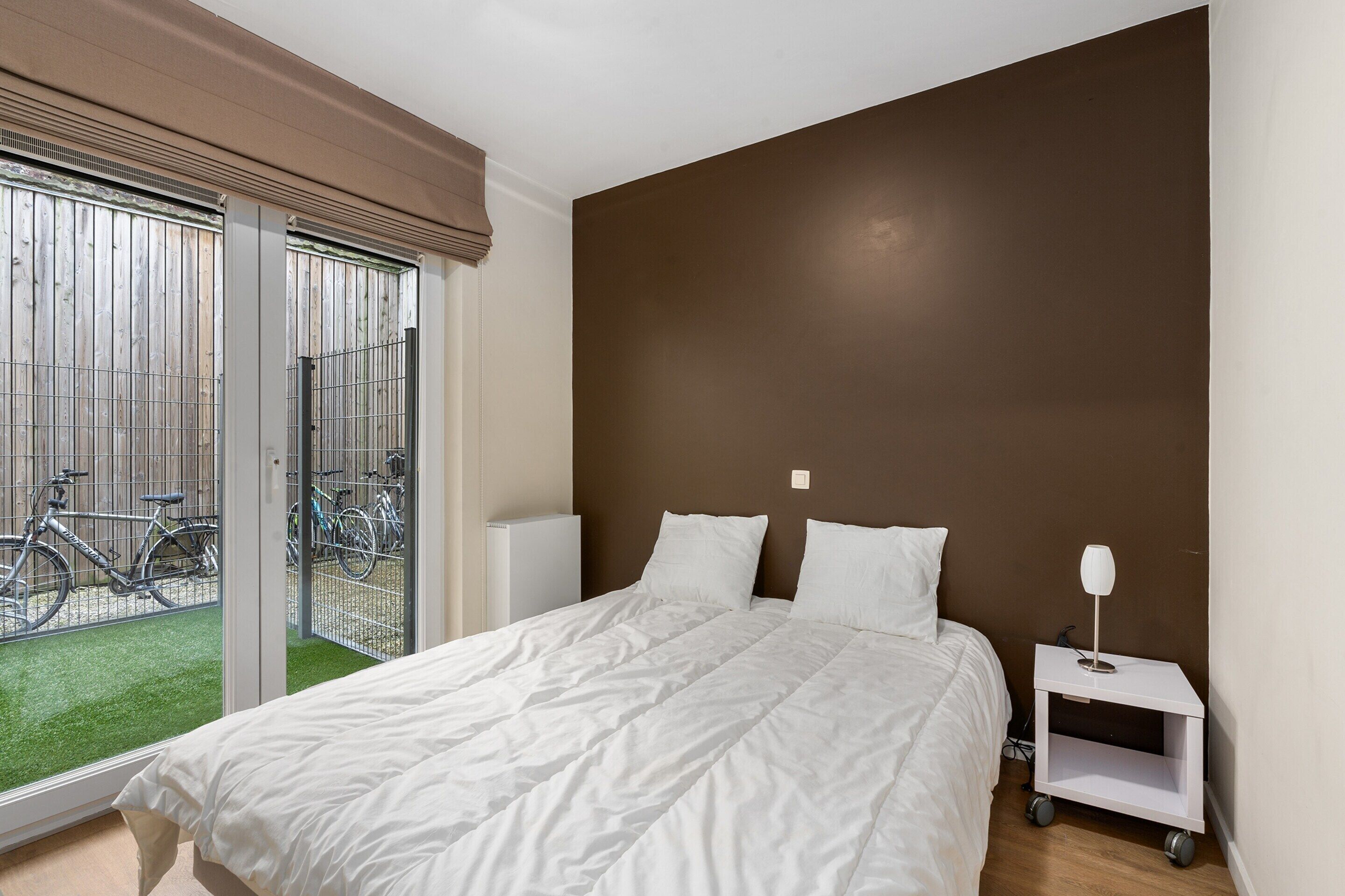 A bedroom with a bed, white pillows, a nightstand with a lamp, and a view of bicycles through a sliding door.