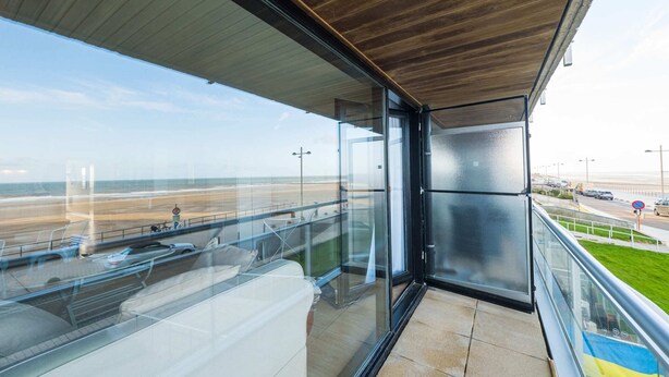 Een modern balkon met glazen omheining, met uitzicht op een strand en een weg.
