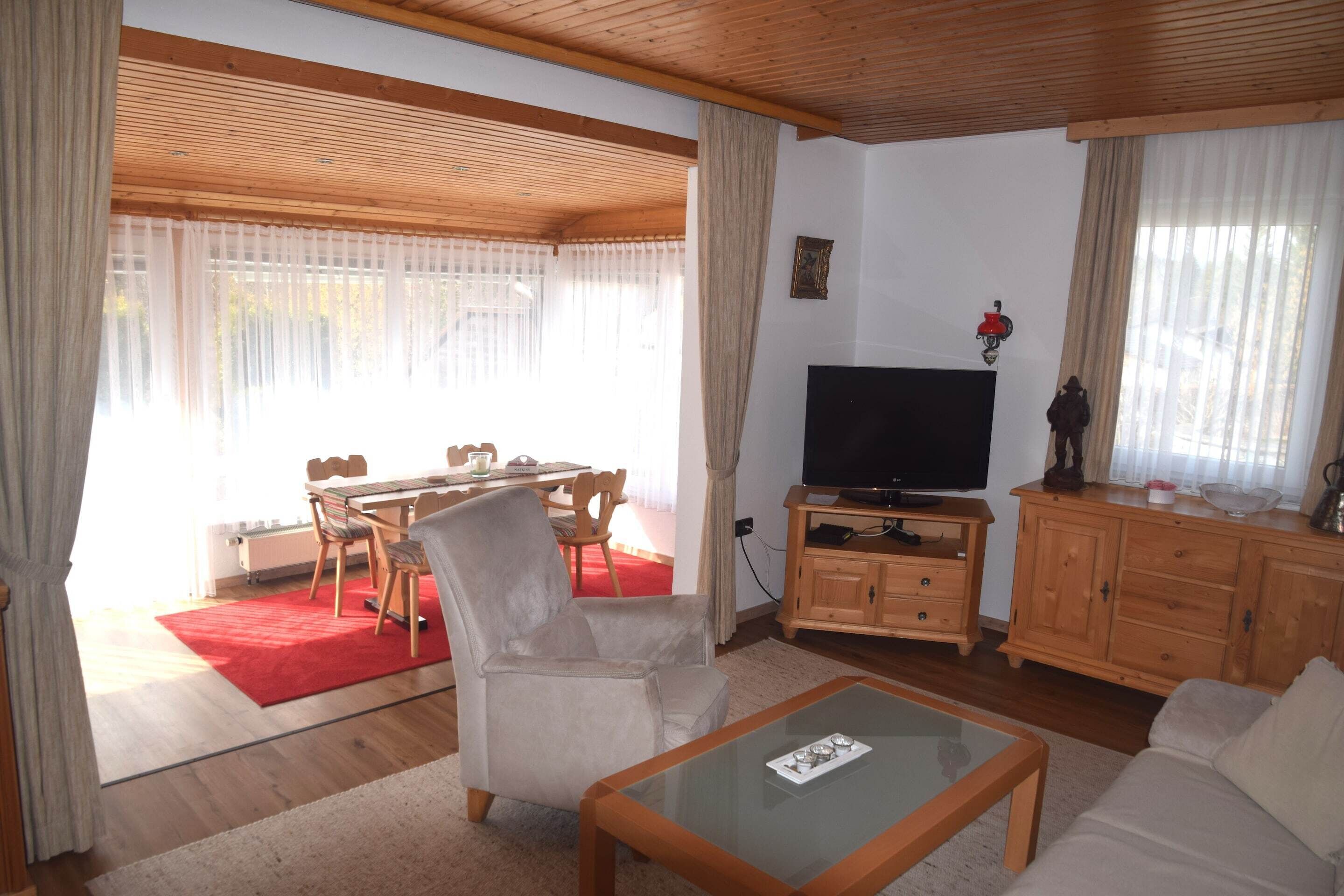 A living room with a television, wooden furniture, a glass coffee table, and a dining area with a red rug.