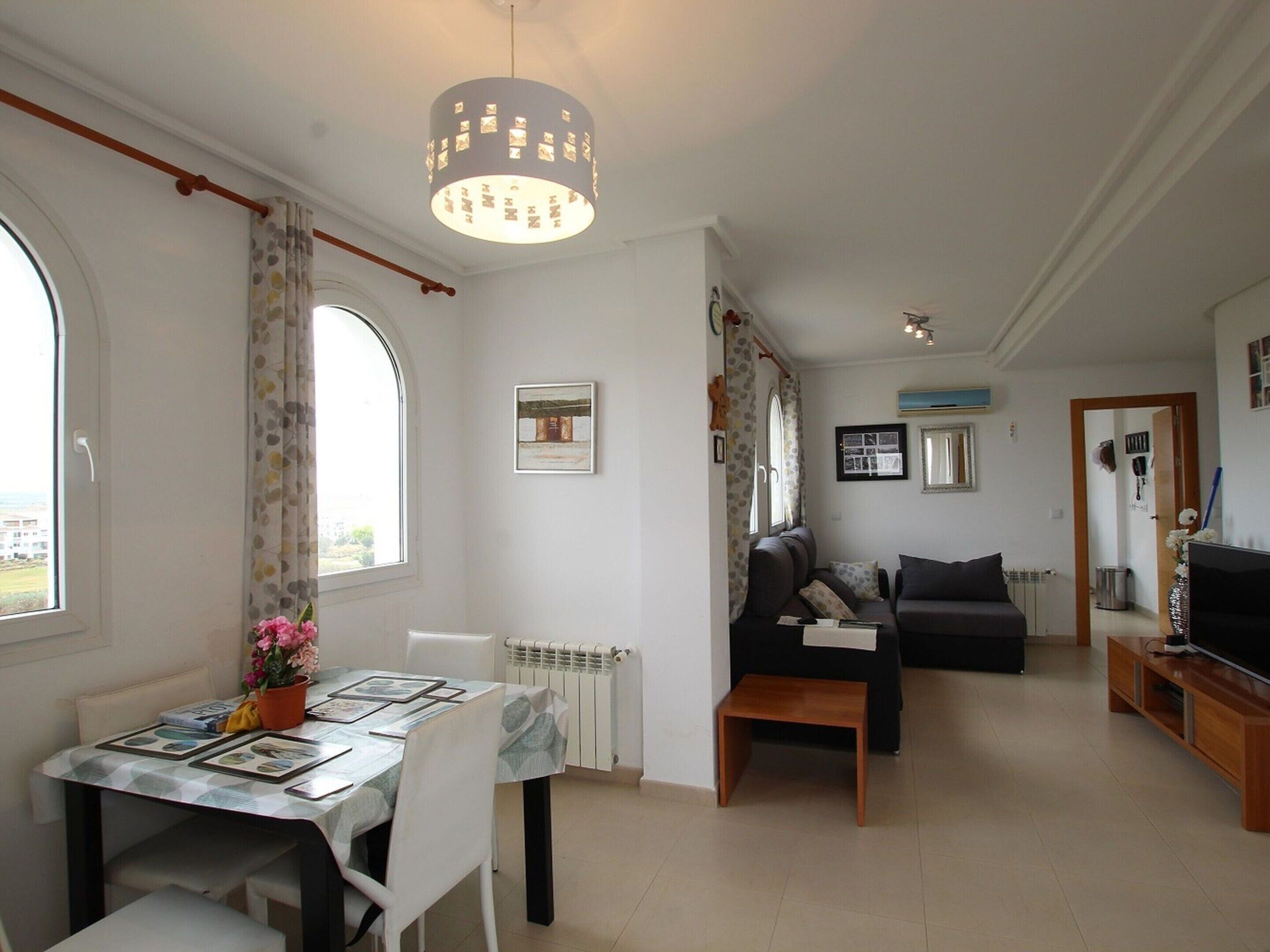 Una sala de estar con mesa de comedor, sofá y mueble para televisor. Se ve una ventana con cortinas, un radiador y una lámpara de techo.