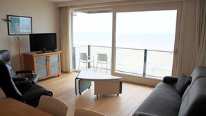 Un salon avec un canapé, un téléviseur posé sur un meuble, une table et un balcon offrant une vue sur la mer.