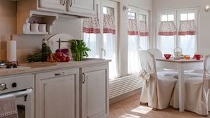 Una cocina con armarios blancos, un comedor con una mesa dispuesta para dos personas y una ventana con cortinas.
