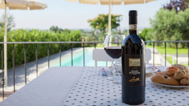Una botella de vino, una copa de vino tinto y un plato de pasteles sobre una mesa al aire libre con mantel a lunares, debajo de una sombrilla blanca.