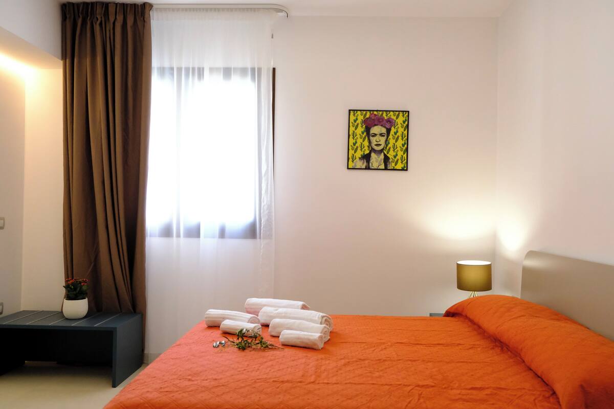 Ein Hotelzimmer mit einem Bett, einem Nachttisch mit Lampe, einem Fenster mit Vorhängen und einem Bild an der Wand.