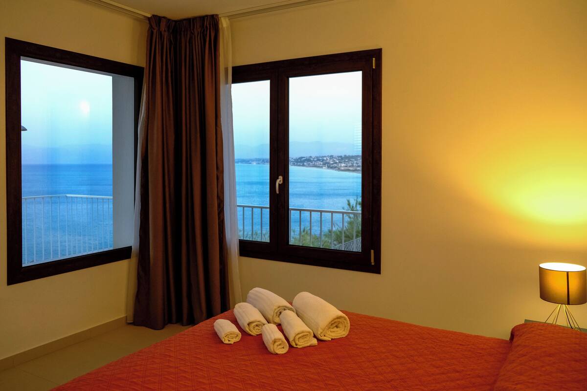 Ein Hotelzimmer mit Bett, einem Vorhangfenster, einer Lampe und Meerblick.