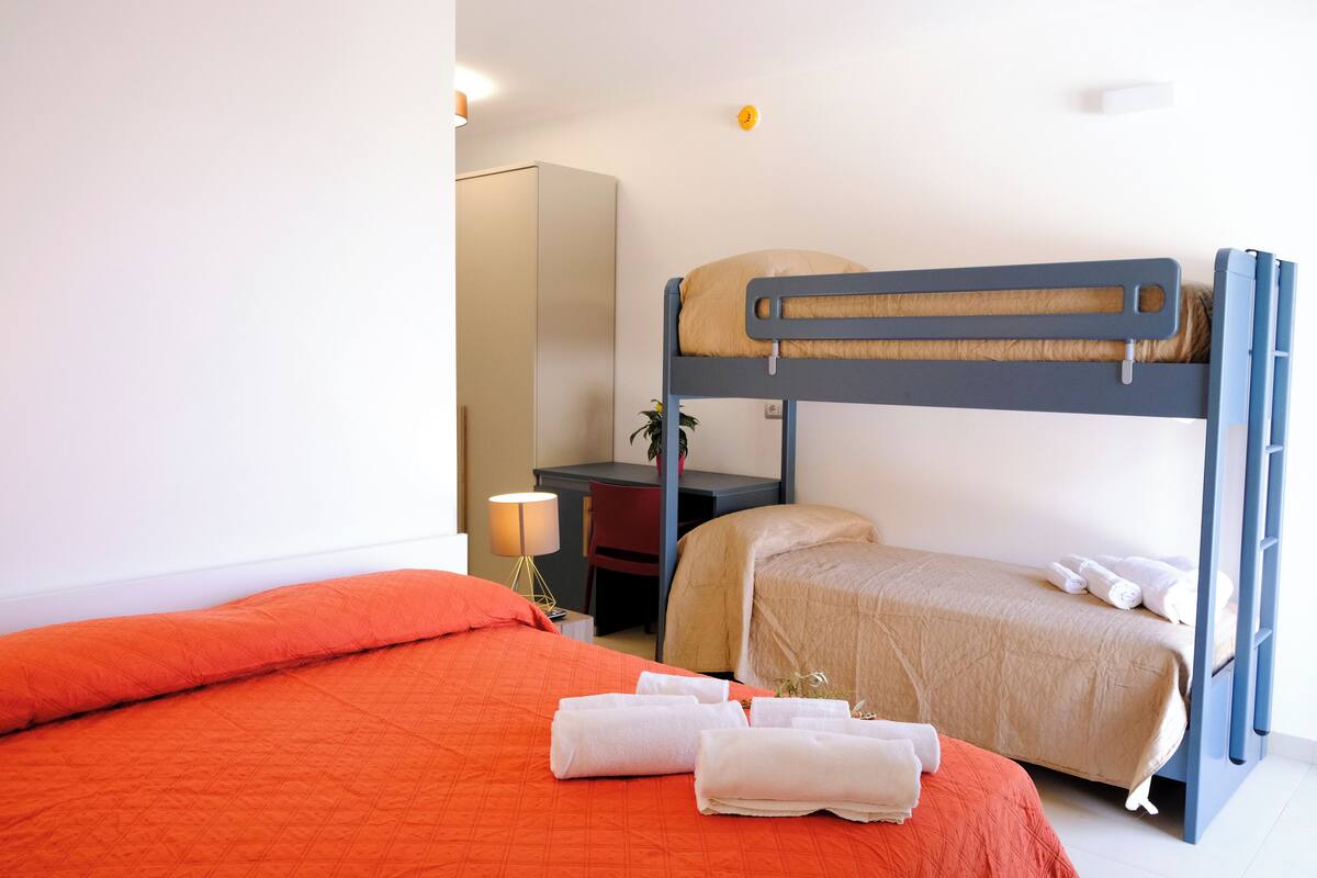 Ein Etagenbett mit Schreibtisch und Stuhl, eine Lampe sowie ein Bett mit orangefarbener Bettwäsche und Handtüchern.