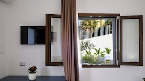 A room with a TV mounted on the wall, a window with a view of a white building and palm trees, and a desk with a red chair and a potted plant. - Sol Y Mar Camere e Appartamenti (Castellammare del Golfo)