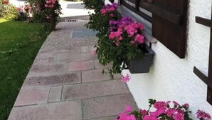 Un sendero conduce a un edificio de fachada blanca y marcos de ventanas oscuros. Hay macetas con flores rosadas en el alféizar de la ventana.