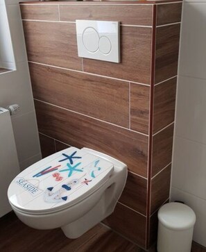 A bathroom with a wooden wall, a white toilet, and a white trash bin. - Lovely Apartment in Hesse (Berkatal)