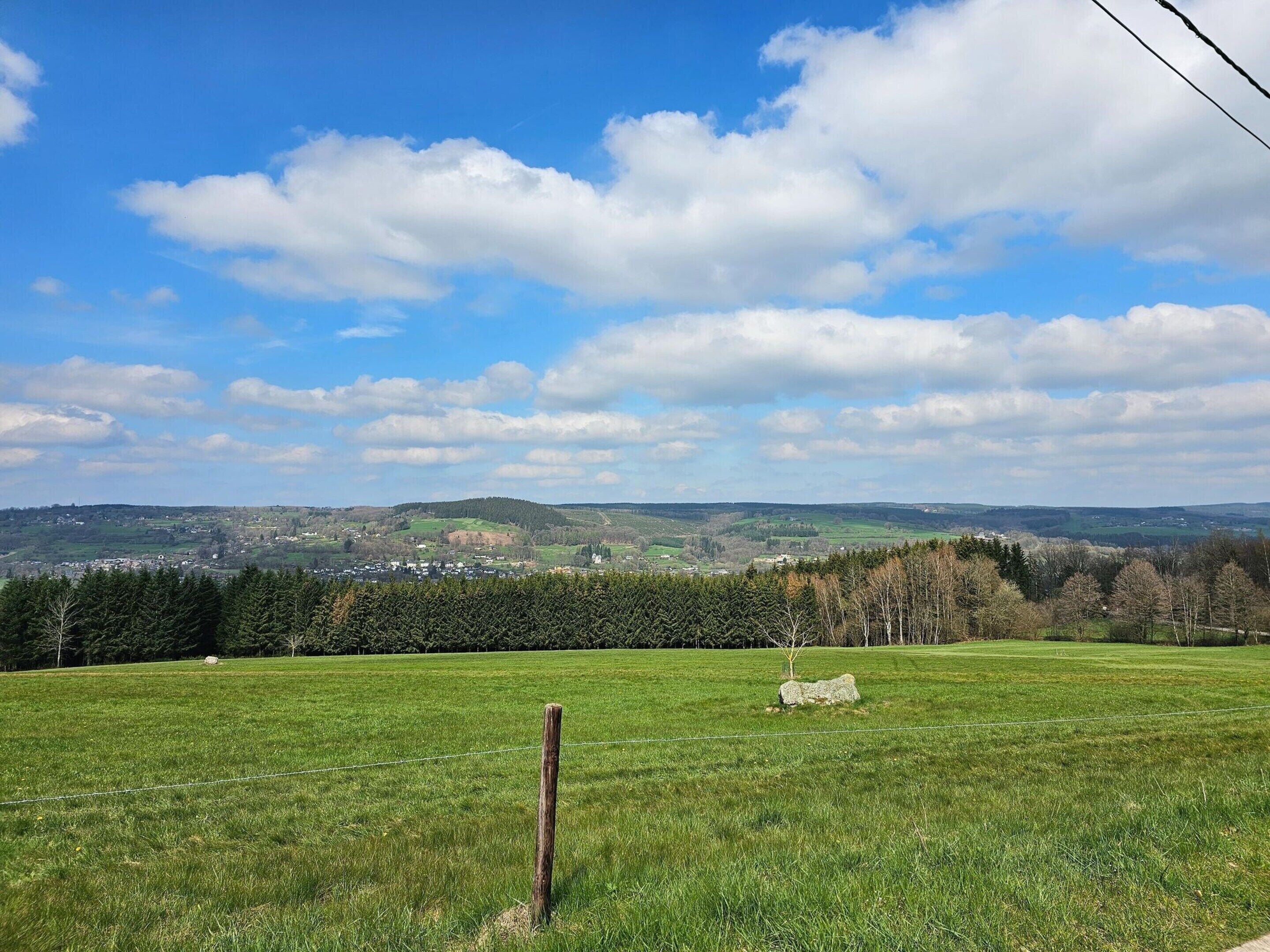 A grassy field with a stone marker and a wooden post, overlooking a town and hills in the distance.