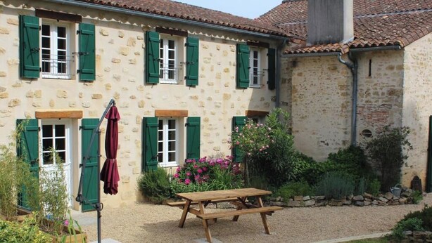 Un bâtiment en pierre avec des volets verts, une table de pique-nique en bois et un chemin de gravier.