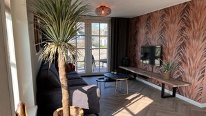 A modern living room with a large potted plant, a sofa, a coffee table, and a TV mounted on the wall. - Villa in Wieringer Near the Wadden Sea (Hippolytushoef)