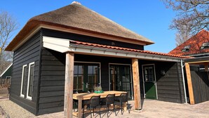 A wooden building with a thatched roof, a covered outdoor seating area with a table and chairs, and a green door. - Villa in Wieringer Near the Wadden Sea (Hippolytushoef)