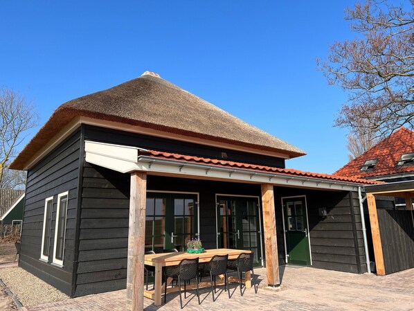 Ein Gebäude aus Holz mit einem Strohdach, einer überdachten Sitzterrasse mit Tisch und Stühlen sowie einer grünen Tür.