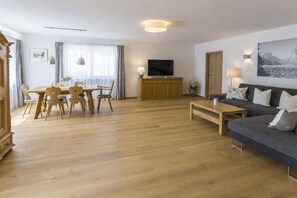 A spacious living room with a wooden floor, a dark grey sofa, a wooden dining table with chairs, a TV on a wooden cabinet, and a painting on the wall. - Marlena Hollental Chalet (Garmisch-Partenkirchen)