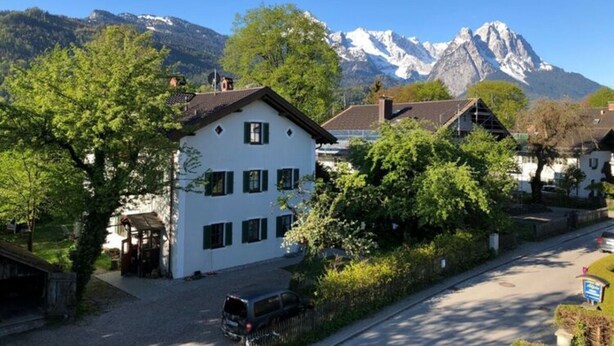 A white building with multiple windows and a sloped roof, surrounded by greenery and a mountain backdrop. - Marlena Hollental Chalet (Garmisch-Partenkirchen)