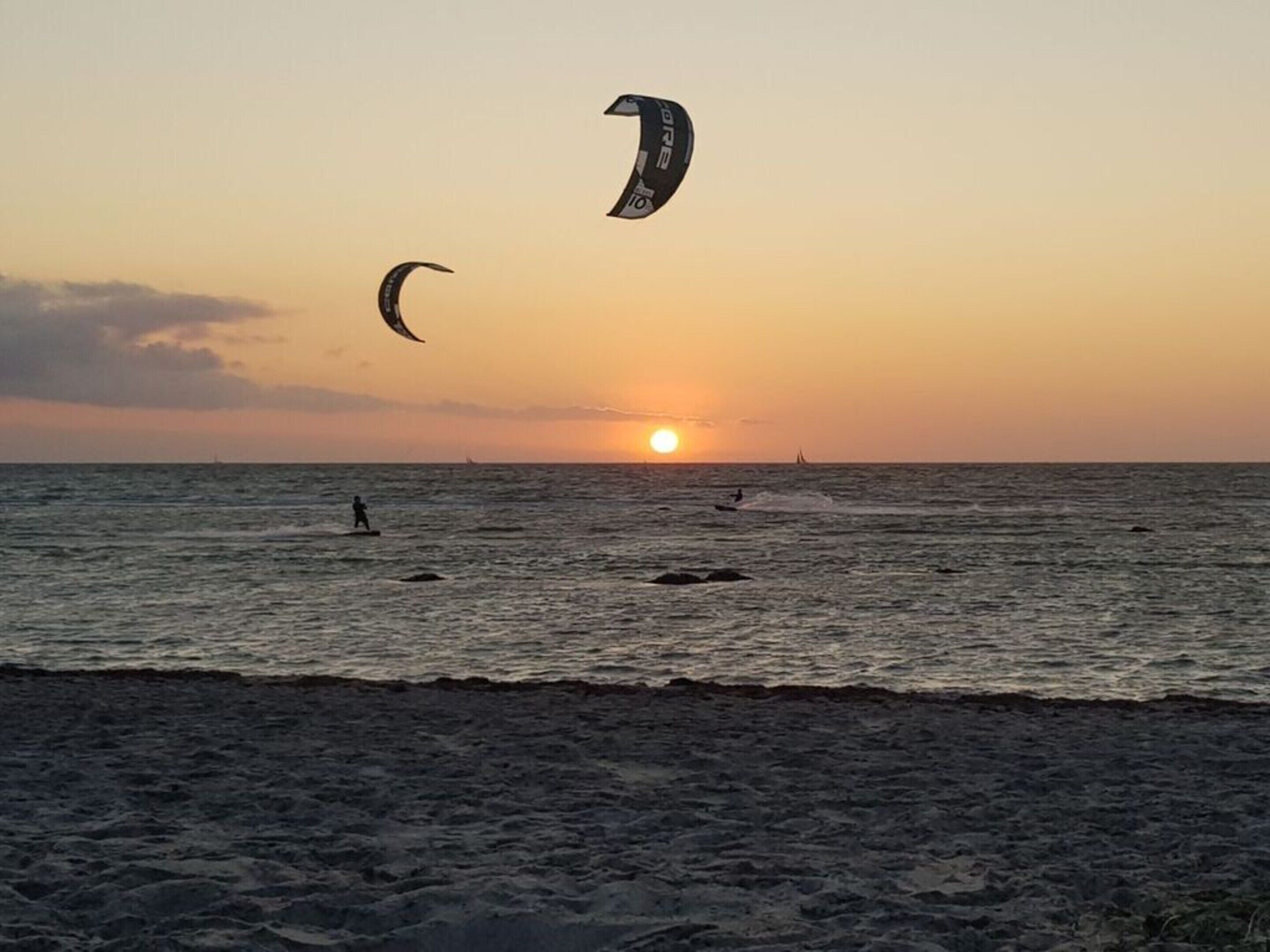 A beach at sunset with a clear sky, calm sea, and a few people in the water.