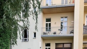 Un bâtiment à deux étages, doté d’un balcon à l’étage supérieur, d’une porte de couleur brun foncé et d’un jardin où poussent des fleurs jaunes et rouges.