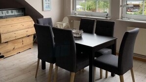 A dining area with a table and chairs, a wooden chest, and a window with a view of trees and buildings. - Holiday Apartment Leah (Toenning)