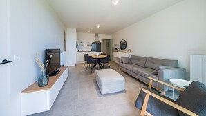 A modern living room with a grey sofa, a small table, and a TV on a stand. - New-build Apartment Near the City Center (Middelkerke)