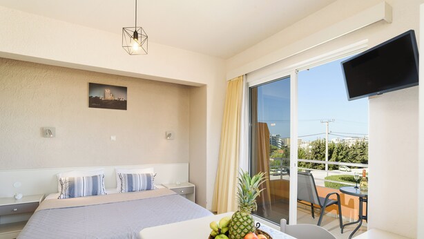 A hotel room with a bed, a table with a fruit bowl, a TV, and a balcony with a view of the city. - Eugenia studios & apartments  (Rhodes)