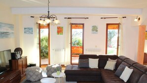 Ein Wohnzimmer mit einem braunen Ecksofa, einem Fernseher und einem Kamin. Zwei Fenster mit Vorhängen, eine Kristallleuchtre und ein kleiner Tisch mit einer Vase sind ebenfalls zu sehen.