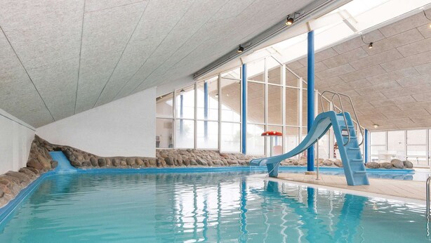 Une piscine intérieure avec un toboggan bleu, des murs en pierre et de grandes fenêtres.