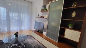 Ein modernes Wohnzimmer mit einem Glastisch, einem Flachbildfernseher, einer Holzvitrine mit Glastüren und einer kleinen Pflanze auf der Fensterbank.