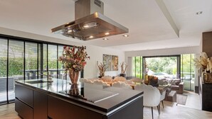 Une cuisine moderne dotée d’un grand îlot central, un coin repas avec une table et des chaises blanches, et un salon avec un canapé et une cheminée.